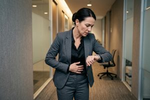 Zakenvrouw van middelbare leeftijd drukt hand op buik in moderne kantoorhal, kijkt bezorgd op horloge